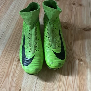 Gröna Nike Mercurial fotbollsskor - Säljer ett par gröna Nike Mercurial fotbollsskor med svart swoosh. Skorna har en strumpliknande passform och är designade för optimal prestanda på planen. Perfekta för den som vill ha både stil och funktion.