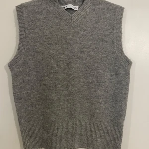 Grå stickad väst från Zara - Säljer en stilren grå stickad väst från Zara. Perfekt för lager-på-lager under kyligare dagar. Västen har en klassisk v-ringad design och ribbade kanter för en snygg passform.