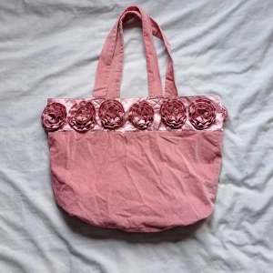 Rosa med blommor - Charmig rosa shoppingväska med dekorativa blommor längs överkanten. Väskan har två handtag och är tillverkad i ett mjukt material. 