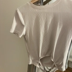 Vit ribbad topp med kedjedetalj - Säljer en stilren vit ribbad topp med korta ärmar och en unik kedjedetalj framtill. Perfekt för att ge din outfit en edgy touch. Passar bra till både jeans och kjol!