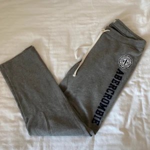 Grå mjukisbyxor från Abercrombie - Säljer ett par grå mjukisbyxor från Abercrombie med vit snörning i midjan och ett tryck längs benet. Perfekta för en avslappnad stil.