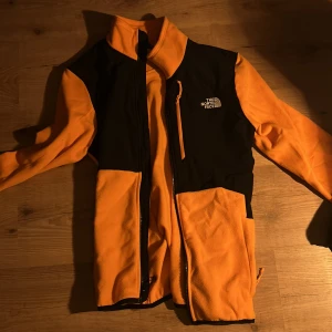 Orange och svart fleecejacka från The North Face - Säljer en snygg orange och svart fleecejacka från The North Face. Jackan har en dragkedja framtill och är långärmad. Perfekt för kyligare dagar och har en stilren design med logotypen på bröstet och ryggen.