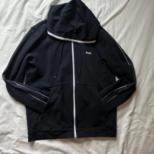 Marinblå hoodie med vita detaljer från Hugo Boss - Säljer en stilren marinblå hoodie från Boss med vita detaljer och dragkedja. Tröjan har en justerbar huva och långa ärmar med subtila logotyper längs sidorna. Nypris 1799:- SEK