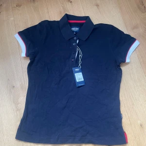 Mörkblå pikétröja från James Harvest - Säljer en snygg mörkblå pikétröja från James Harvest med röda och vita detaljer. Tröjan har korta ärmar och en klassisk krage med knappar framtill. Perfekt för en sportig look!