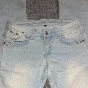 Ljusblå jeans med strassdetaljer - Snygga ljusblå jeans med strassdetaljer på fram- och bakfickorna. De har en klassisk femficksdesign och en knappgylf. Perfekta för att ge en glittrig touch till din outfit.