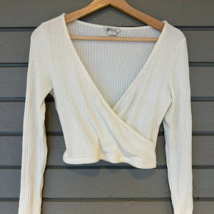 Vit ribbad omlottopp från Monki - Säljer en stilren vit ribbad omlottopp från Monki. Toppen har en v-ringad design och långa ärmar. Den är croppad och passar bra till både jeans och kjol.