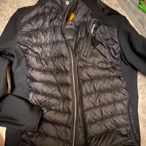 Svart jacka från Parajumpers - Säljer en svart jacka från Parajumpers med quiltad design och dragkedja. Jackan har en cool patch på ärmen och är perfekt för kyligare dagar. Den har en stilren look med praktiska fickor.