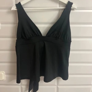 Svart topp med knytdetalj på baksidan av tröjan från Nelly - Snygg svart topp från Nelly med djup v-ringning och knytband i midjan. Perfekt för en stilren look. Tillverkad i ett mjukt och bekvämt material. Aldrig använd och aldrig testad. Köpte från Nelly. Storlek:XL. Säljer för 175 kronor 