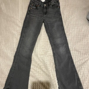 Grå jeans från Gina Tricot - Snygga grå jeans från Gina Tricot i storlek 152. De har en klassisk femficksdesign med knapp och dragkedja framtill. Byxorna har en bootcut-stil som ger en trendig look. Byxorna har en liten fläck på baksidan