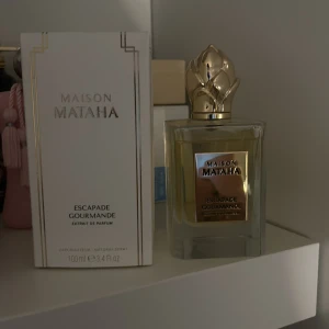Maison Mataha Escapade Gourmande Parfym - Lyxig parfym från Maison Mataha, Escapade Gourmande, i en elegant flaska med guldfärgad detalj och dekorativ kork. Förpackningen är stilren med guldaccenter. Perfekt för den som söker en exklusiv doftupplevelse.