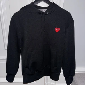 Svart hoodie från Comme des Garçons - Säljer en stilren svart hoodie från Comme des Garçons med det ikoniska röda hjärtat på bröstet. Tröjan har en klassisk passform med huva och dragsko. Perfekt för en avslappnad look. Använd fåtal gånger.