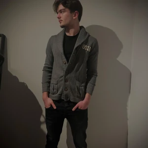 Grå kofta med knappar - Snygg grå kofta med knappar och broderad detalj på bröstet. Perfekt för en stilren look. Koftan har långa ärmar och ribbade muddar för extra komfort.