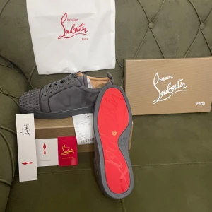 Grå sneakers från Christian Louboutin - Snygga grå sneakers från Christian Louboutin med den ikoniska röda sulan. Skorna har en cool design med nitar på tån och är tillverkade i mocka. Perfekta för dig som vill sticka ut med stil.