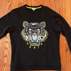 Svart sweatshirt från Kenzo med tigerbroderi - Snygg svart sweatshirt från Kenzo med ett stort, färgglatt tigerbroderi på framsidan. Tröjan har långa ärmar och en rund halsringning. Perfekt för en stilren och trendig look.