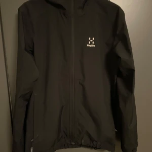 Svart vindjacka från Haglöfs - Snygg svart vindjacka från Haglöfs med dragkedja och huva. Perfekt för blåsiga och regniga dagar. Jackan har en stilren design med Haglöfs logga på bröstet. Den är knappt använd och har inga täcken på användning. Byten är intressant. Skriv om ni undrar något!