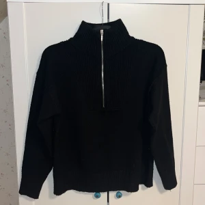 Svart stickad tröja med dragkedja  - Säljer en stilren svart stickad tröja från KappAhl med dragkedja framtill. Tröjan har en hög krage och långa ärmar, perfekt för kyligare dagar. Den ribbade designen ger en snygg och bekväm passform.