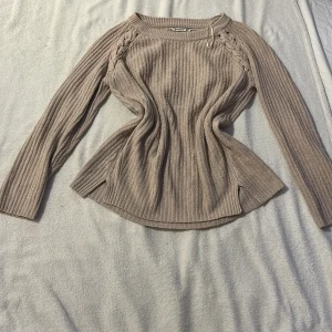 Beige ribbad tröja från strandivarius - Säljer en beige ribbad tröja från Strandivarius med flätade detaljer vid axlarna. Tröjan har långa ärmar och en rund halsringning. Perfekt för kyligare dagar. Passar bra till jeans eller kjol. 🌟En liten fläck på ens armen som ej syns så mycket och borde gå bort med rätt tvätt