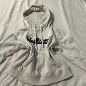 Vit hoodie från Hollister - Säljer en vit hoodie från Hollister med broderad logga i svart på bröstet. Den har en klassisk känguruficka och justerbar huva med dragsko. Perfekt för en avslappnad stil.