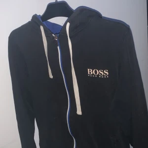 Svart hoodie från Hugo Boss - Säljer en stilren svart hoodie från Hugo Boss med vit dragkedja och snören. Den har en blå insida i huvan och ett klassiskt BOSS-tryck på bröstet. Perfekt för en avslappnad look. Passar S/XS