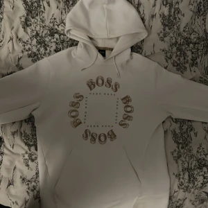 Vit hoodie från Hugo Boss - Säljer en stilren vit hoodie från Hugo Boss med ett cirkulärt BOSS-tryck på framsidan. Hoodien har en klassisk design med en stor ficka framtill och en bekväm huva. Perfekt för en avslappnad look.