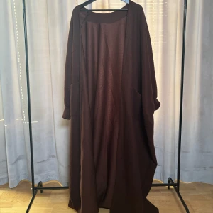 Brun lång kimono - Säljer en elegant brun lång kimono med en avslappnad passform. I storlek one size. Den har långa ärmar och praktiska fickor framtill. Perfekt för kyligare dagar och ger en stilren look. 