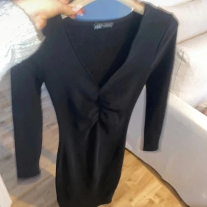Svart klänning från Zara storlek S - Snygg svart klänning från Zara med v-ringning och långärmad design. Klänningen har en åtsittande passform och en elegant rynkdetalj framtill. Perfekt för en stilren look. Storlek S