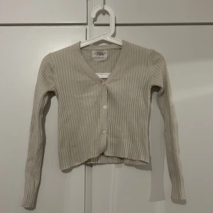 Beige ribbad kofta från Zara - Säljer en stilren beige ribbad kofta från Zara i storlek 13-14. Koftan har en v-ringad design med knappar framtill och långa ärmar.
