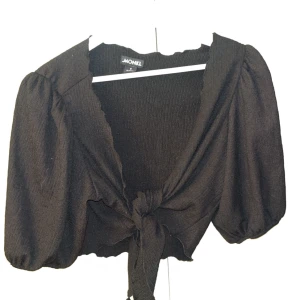 Svart knytblus från Monki - Snygg svart blus från Monki med knytning framtill och puffiga ärmar. Den har en croppad passform och är tillverkad i ett lätt och bekvämt material. Perfekt för en stilren look.