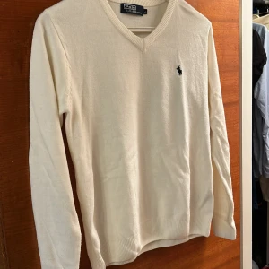 Vit tröja från Polo Ralph Lauren - Säljer en stilren vit tröja från Polo Ralph Lauren med v-ringning och det klassiska logotypbroderiet på bröstet. Tröjan har långa ärmar och ribbade kanter vid ärmslut och nederkant. Perfekt för en elegant och avslappnad look.