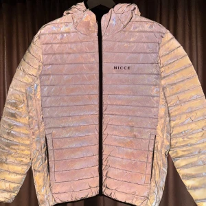 Reflekterande jacka från Nicce - Säljer en reflekterande jacka från Nicce i storlek M. Jackan har en cool, skimrande yta som ändrar färg beroende på ljuset. Den är quiltad med dragkedja och har långa ärmar. Perfekt för att sticka ut i mängden!