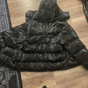 Svart glansig dunjacka från Trapstar  - Säljer en svart glansig dunjacka från Trapstar med huva och dragkedja. Jackan har ett märke på bröstet och text på ryggen. Perfekt för kyliga dagar.