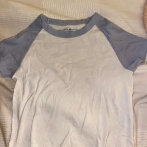 Vit och blå t-shirt (lite croppad?) - Använd max en gång. Säljer en stilren t-shirt med vit bas och ljusblå ärmar. Perfekt för en avslappnad look. T-shirten har korta ärmar och en rund halsringning. Priset kan diskuteras 