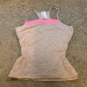 Grå linnetopp med rosa spetsdetalj från H&M - Snygg grå linnetopp från H&M med smala axelband och en söt rosa spetsdetalj vid bysten. Perfekt för varma dagar eller som ett lager under en tröja. Säljer även det här linnet med ett par matchande pyjamas shorts, om man vill hellre ha ett set. Storlek XS. 