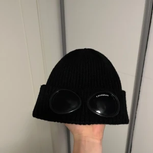 C.P. Company mössa - Snygg svart ribbad mössa från C.P. Company med unika glasögondetaljer framtill. Den är inte använd mycket så bra i skick, storlek ONE SIZE.