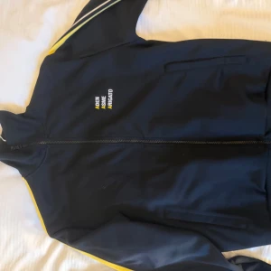 Svart set från Axel Arigato x aden x asme - Snygg svart tracksuit från Axel Arigato med dragkedja och gula detaljer. Koftan har vita och gula ränder längs ärmarna och text på bröstet. Perfekt för en sportig look.