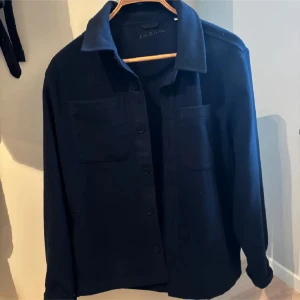 Mörkblå overshirt från donce de la sense - Snygg mörkblå overshirt från donce de la sense med knappar framtill och två bröstfickor. Perfekt för lager-på-lager-stil. Den har en klassisk krage och långa ärmar, vilket gör den idealisk för kyligare dagar. Kan gå ner i pris i snabba affärer
