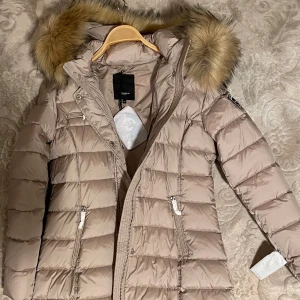 Beige dunjacka från Rockandblue - Snygg beige dunjacka från Rockandblue med pälskantad huva. Jackan har en quiltad design och dragkedja framtill. Perfekt för kyliga dagar med sin långa ärm och stilrena look. Använt en gång kort stund och tog av etiketter.