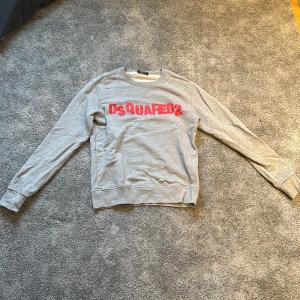 Grå sweatshirt från Dsquared2 - Säljer en grå sweatshirt från Dsquared2 med röd logga på bröstet. Tröjan är tillverkad i 100% bomull och har långa ärmar. Nypris ligger över 2000 kr