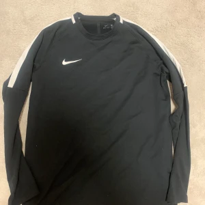 Svart långärmad tröja från Nike - Säljer en svart långärmad tröja från Nike med vita detaljer på ärmarna och en vit Nike-logga på bröstet. Perfekt för sportiga aktiviteter eller en avslappnad stil. Tröjan har en bekväm passform och är tillverkad i ett mjukt material.