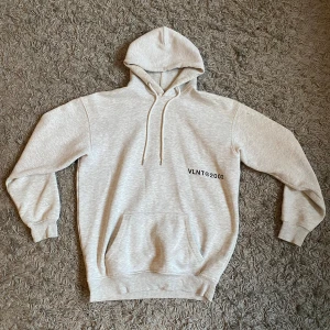 Grå hoodie från Vailent - Säljer en stilren grå hoodie från Vailent med tryck 'VLNT©2002' på bröstet. Helt nytt skick, då jag bara testat den! 250kr
