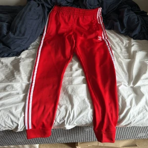 Röda träningsbyxor från Adidas - Snygga röda träningsbyxor från Adidas med klassiska vita ränder längs sidorna. Byxorna har en elastisk midja och är perfekta för sportiga aktiviteter. Tillverkade med Parley Ocean Plastic för en hållbar touch.