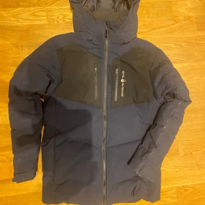 Mörkblå dunjacka från Sail Racing strl 160 - Snygg mörkblå dunjacka från Sail Racing med svarta detaljer som är inköpt på Kids Brandstore. Har dock lite skav på ena ärmen (se separat bild) och några småstänk på vä bröst. Har aldrig tvättat jackan så det kan nog försvinna när man gör det. 