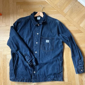 Mörkblå jeansjacka från Lee - Säljer en snygg mörkblå jeansjacka från Lee med knappar framtill och en bröstficka med logotyp. Jackan har långa ärmar och en klassisk krage. Perfekt för en avslappnad stil.