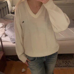 Vit tröja från Lacoste - Säljer en stilren vit tröja från Lacoste med v-ringning och ribbade detaljer. Tröjan har långa ärmar och den klassiska krokodilloggan på bröstet. 