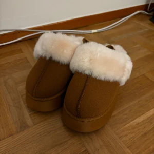 Bruna tofflor med vit päls - Mysiga bruna tofflor med fluffig vit päls runt kanten. Perfekta för kalla dagar inomhus. De har en stabil sula och en bekväm passform. Perfekta för att hålla fötterna varma och sköna under vintern. De är helt nya, det står storlek 40 på de men de passar som 39. 