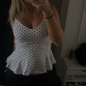 Vit prickig topp med volang - Säljer en superfin vit topp med svarta prickar och volangdetalj i nederkant. Toppen är ärmlös och har en tight passform. Perfekt för sommarens alla fester! 🖤