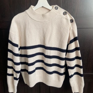 Stickad tröja Bondelid - Stickad tröja med knappar på axeln från Bondelid. Off-white med mörkblå ränder. Mycket bra skick. Normal i storlek.   Modell: Angelicka stripe knit