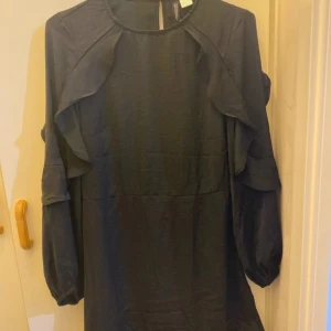 Svart klänning från H&M - Säljer en elegant svart klänning från H&M i storlek 38. Klänningen har långa ärmar och fina volangdetaljer längs axlarna. Perfekt för höst- och vinterfester. Materialet känns mjukt och bekvämt. Passar både till vardags och festligare tillfällen.