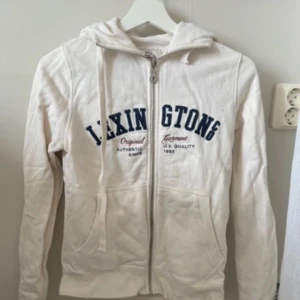 Vit hoodie från Lexington - Säljer en vit hoodie från Lexington med dragkedja och tryck i blått på framsidan. Den har en mysig huva med snören och långa ärmar. Perfekt för en avslappnad stil eller kyliga dagar. Hör av dig vid intresse!