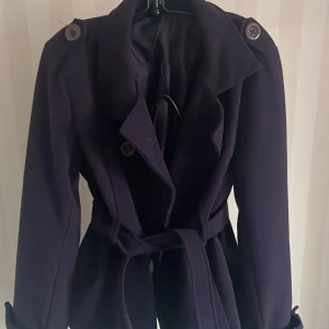 Mörklila trenchcoat / jacka med bälte - Snygg mörklila trenchcoat i nyskick. Den har långa ärmar och ett bälte i midjan för en elegant look. Perfekt för höst och vår. Knapparna på axlarna och ärmsluten ger en extra touch av stil. Passar både till vardags och fest!
